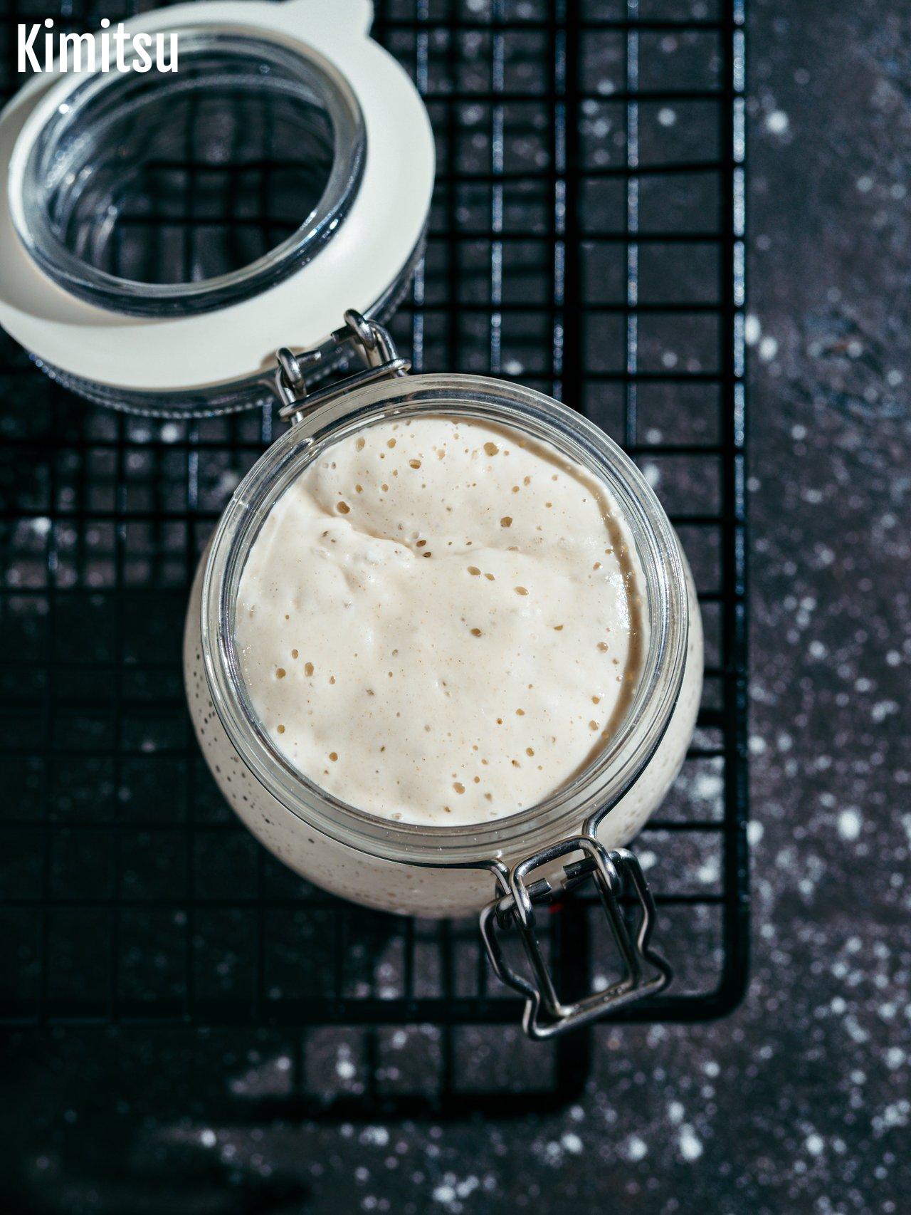 Pot en verre avec un sourdough starter en fermentation, bulles visibles sur pâte hydratée et lisse.